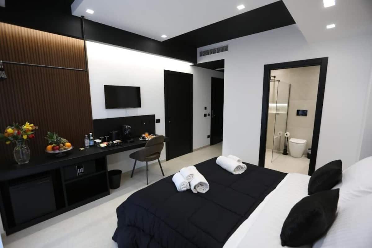 A modern hotel room with a bed, a desk, a chair, a TV, and a bathroom visible through an open door.