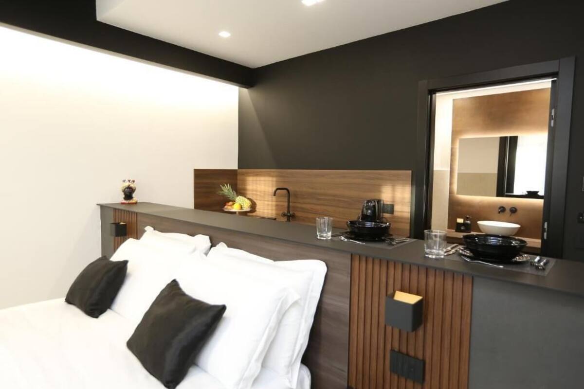 A modern hotel room with a bed, a wooden vanity, a mirror, and a small kitchenette.