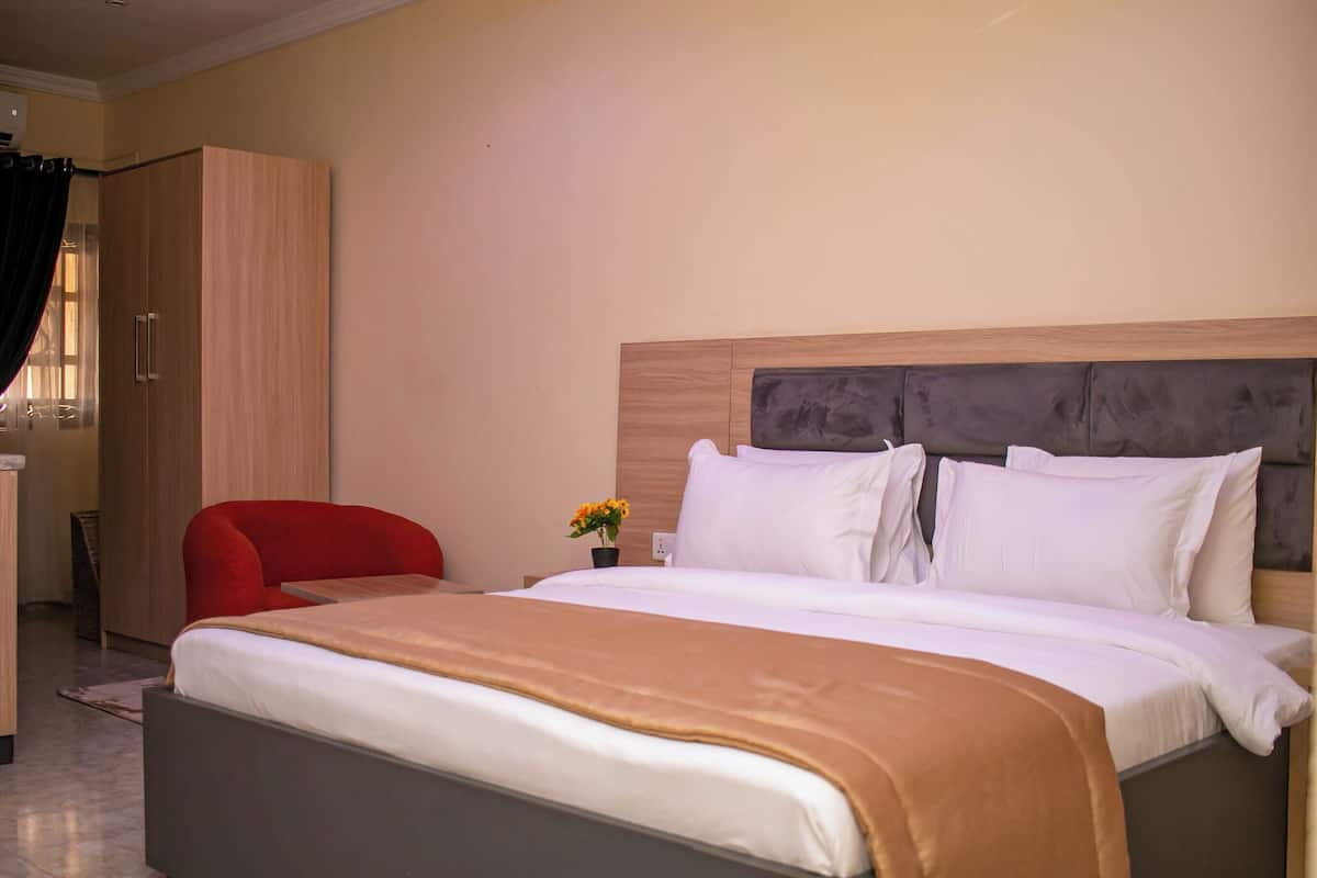 Una habitación de hotel con una cama, una silla roja, un armario de madera y una mesita con una flor.