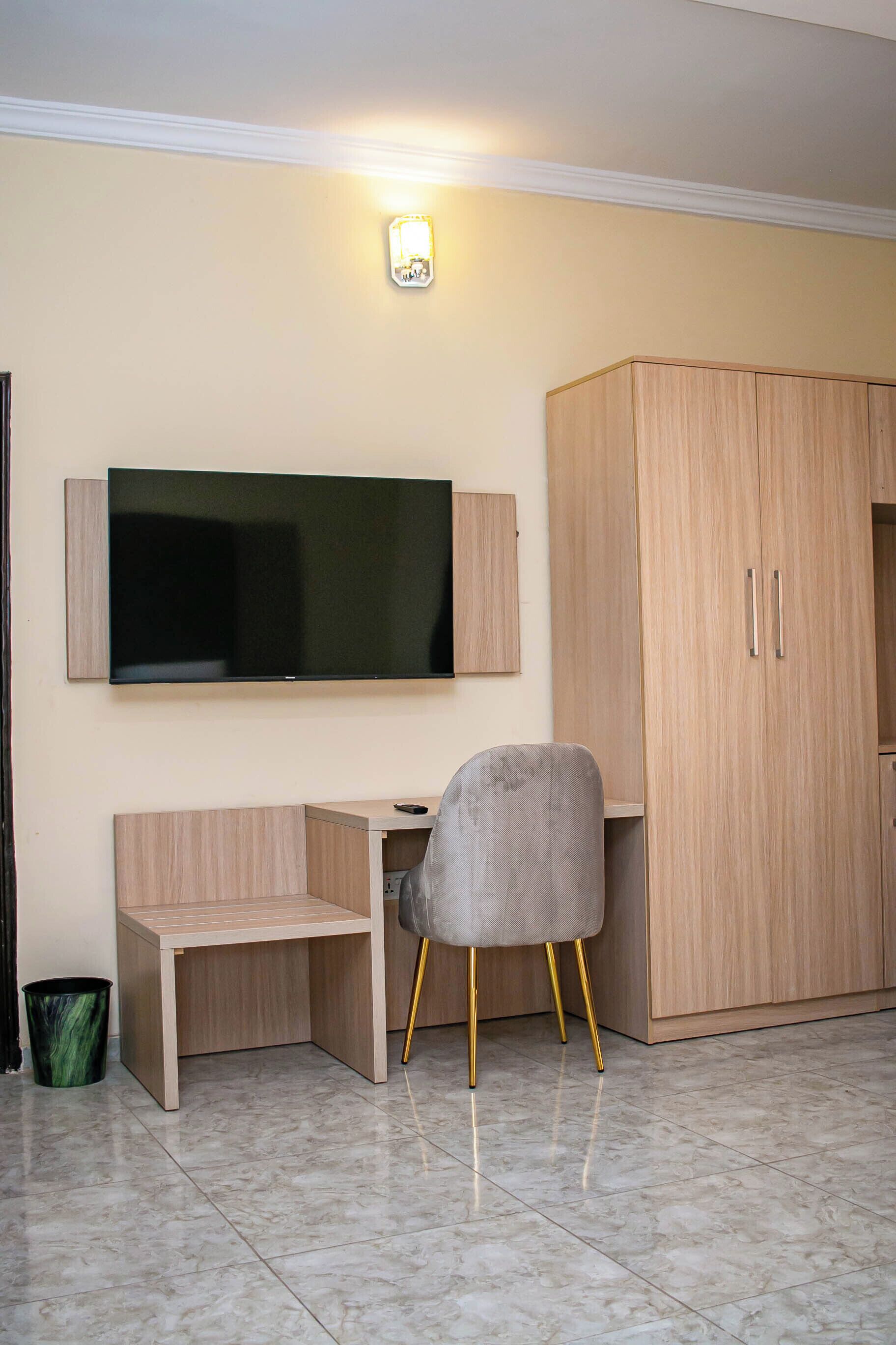 Una habitación con una televisión de pantalla plana montada en la pared, un escritorio de madera, una silla gris con patas doradas y un armario grande.