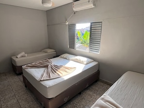 A room with two beds, a window with blinds, an air conditioning unit, and a tiled floor.