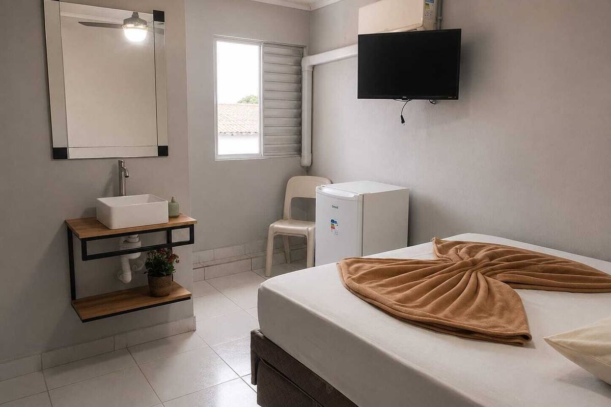 Un dormitorio con cama, televisor, lavamanos, silla y ventana.