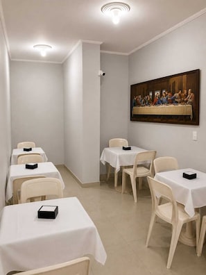 A dining area with tables and chairs, a ceiling light, and a painting on the wall. - Pousada Aviação (Praia Grande)