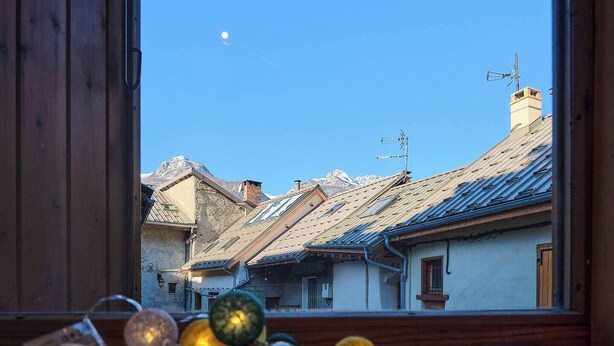 Blick durch ein Fenster auf ein Gebäude mit einem geneigten Dach, sichtbaren Schornsteinen und einem klaren Himmel mit einem Mond.