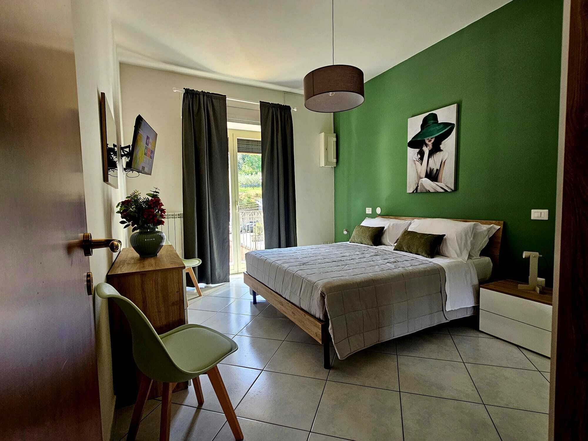 Une chambre à coucher avec un lit, un bureau, une chaise, un mur vert et une fenêtre avec des rideaux.