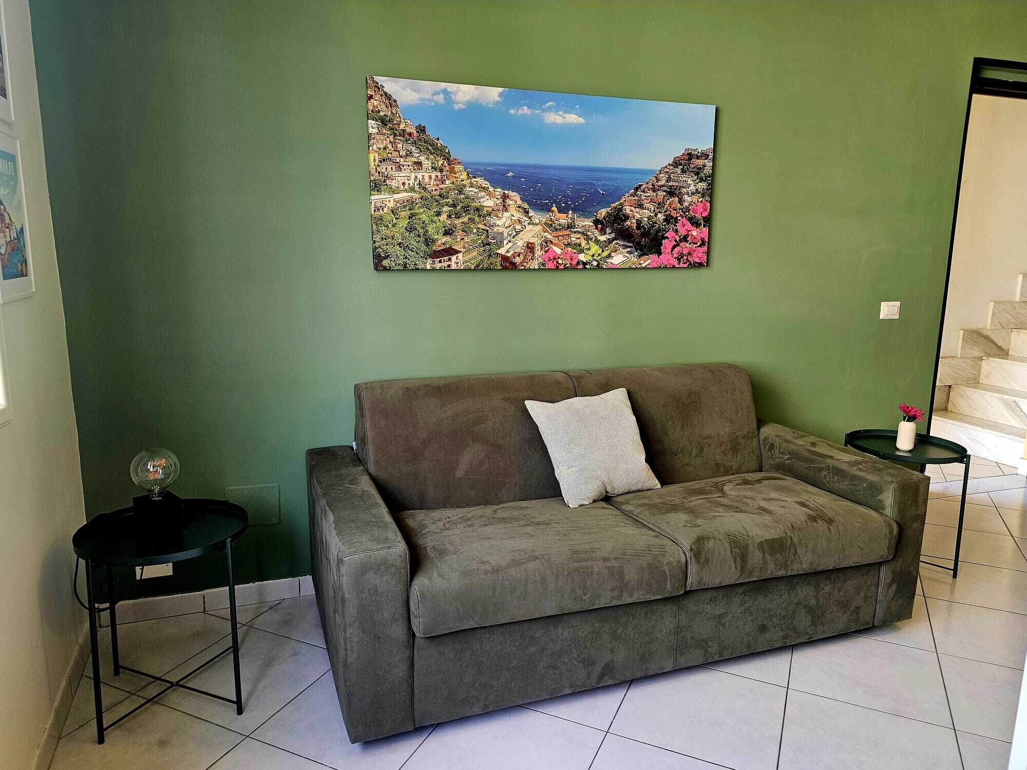 Un salón con una pared verde, un sofá gris, una mesita auxiliar negra pequeña, un cuadro enmarco con un paisaje costero, y una escalera al fondo.