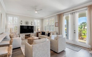 Ein geräumiges Wohnzimmer mit Flachbildfernseher, bequemen Sitzmöglichkeiten, großen Fenstern und einem Deckenventilator.