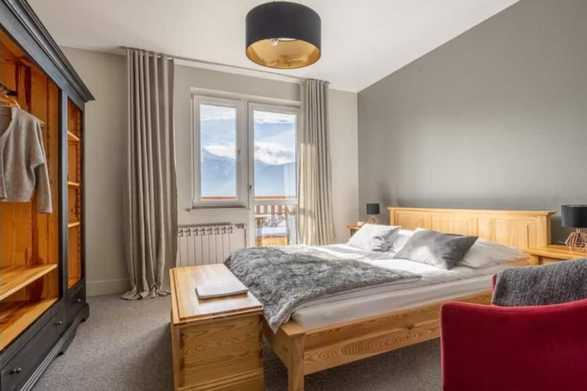 Un dormitorio con una cama, una mesita de noche de madera, una silla roja, un armario y una ventana con cortinas.
