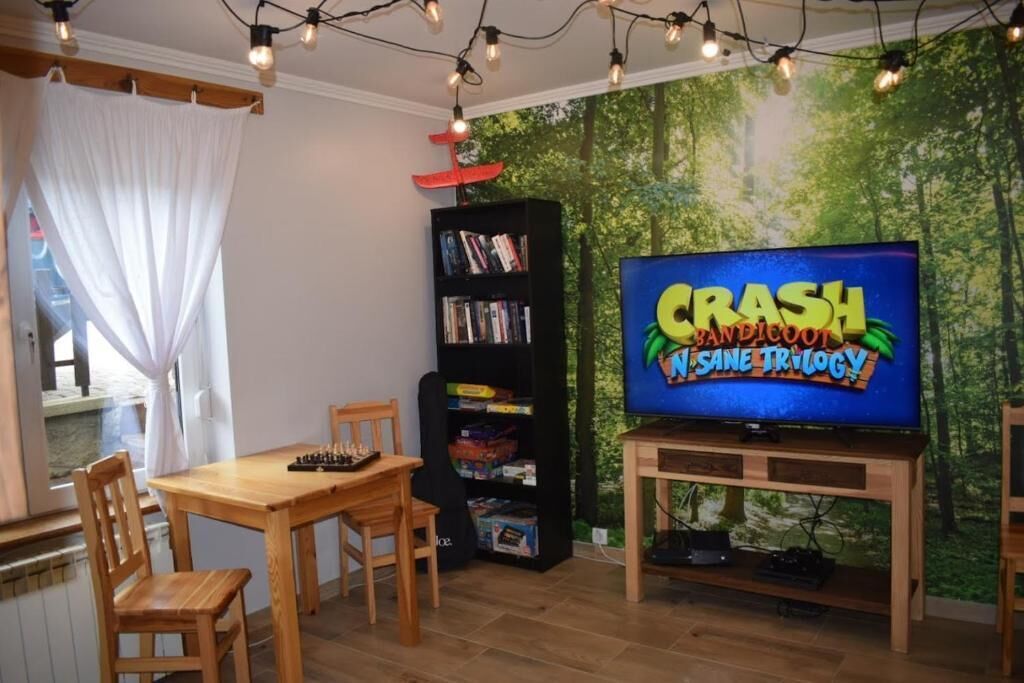 Una stanza con una televisione che mostra 'CRASH MANSION 1 SANE TRILOGY', una libreria, un tavolo in legno e delle sedie.