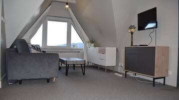 Een moderne loft slaapkamer met een schuine plafond, een groot raam, een grijze bank, een kleine tafel, een televisie op een houten standaard, en een witte dressoir.