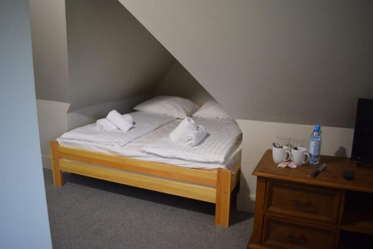Una cama individual con estructura de madera, una mesita de noche con una botella de agua y un televisor sobre un mueble bajo.