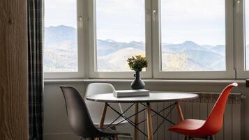 A room with a table, two chairs, a vase with flowers, and a view of mountains through large windows.