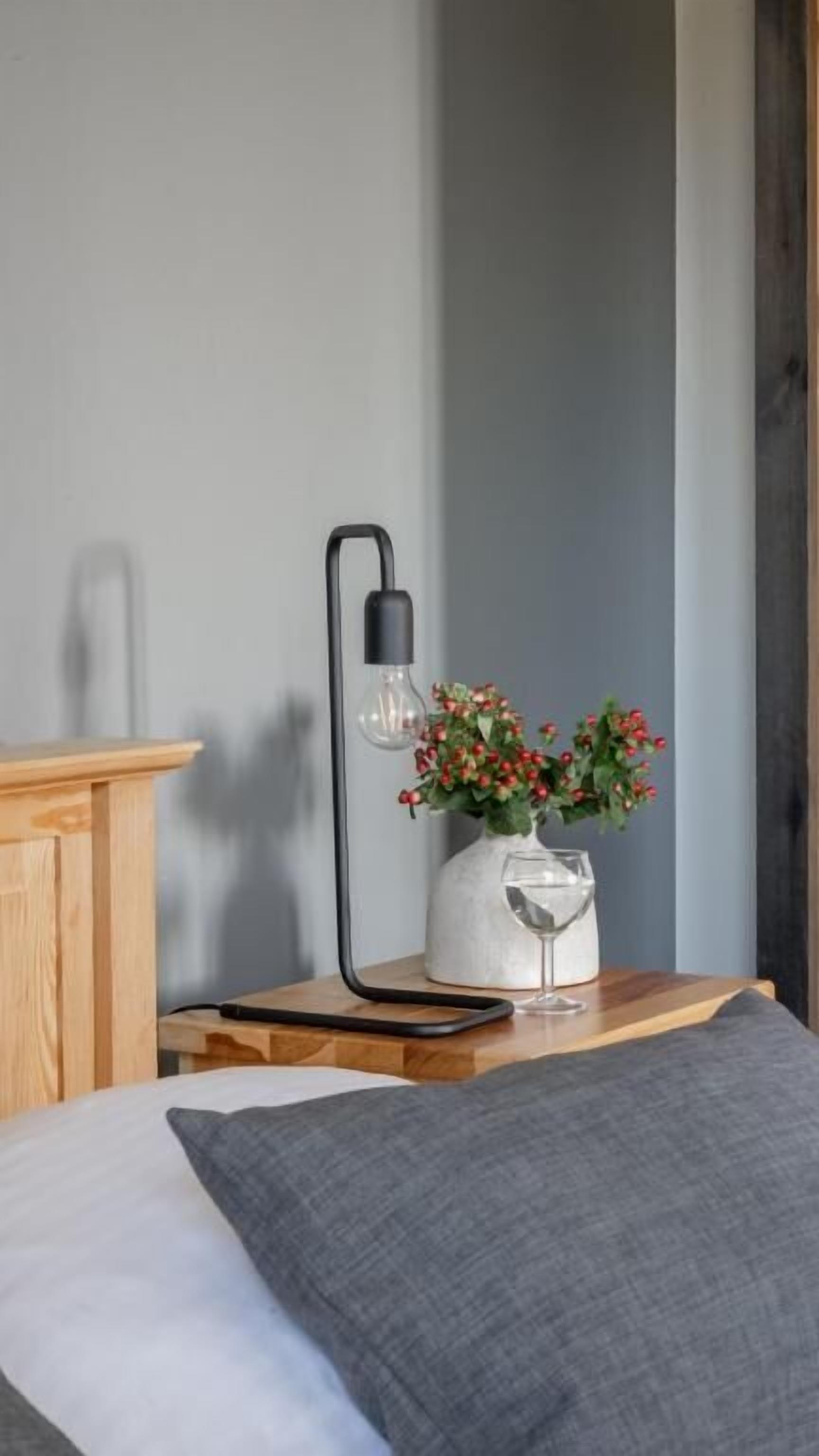 Un lit avec un oreiller gris, une table de chevet en bois sur laquelle se trouve un vase rempli de baies rouges, et une lampe noire.