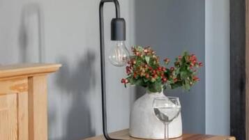 Un lit avec un oreiller gris, une table de chevet en bois sur laquelle se trouve un vase rempli de baies rouges, et une lampe noire.