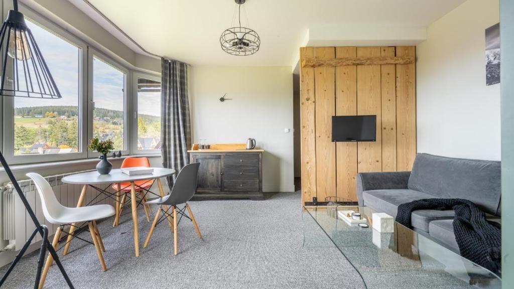 Un salon moderne avec un canapé, une table à manger et des chaises, un téléviseur fixé à un mur en bois, et une vue dégagée grâce à de grandes fenêtres.