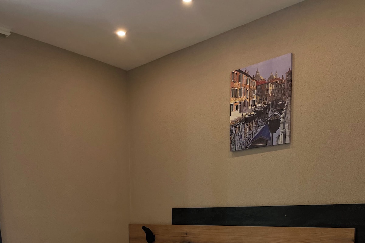 Una cama bien hecha con sábanas y almohadas blancas, cabecera de madera y un cuadro colgado en la pared que muestra una escena callejera.