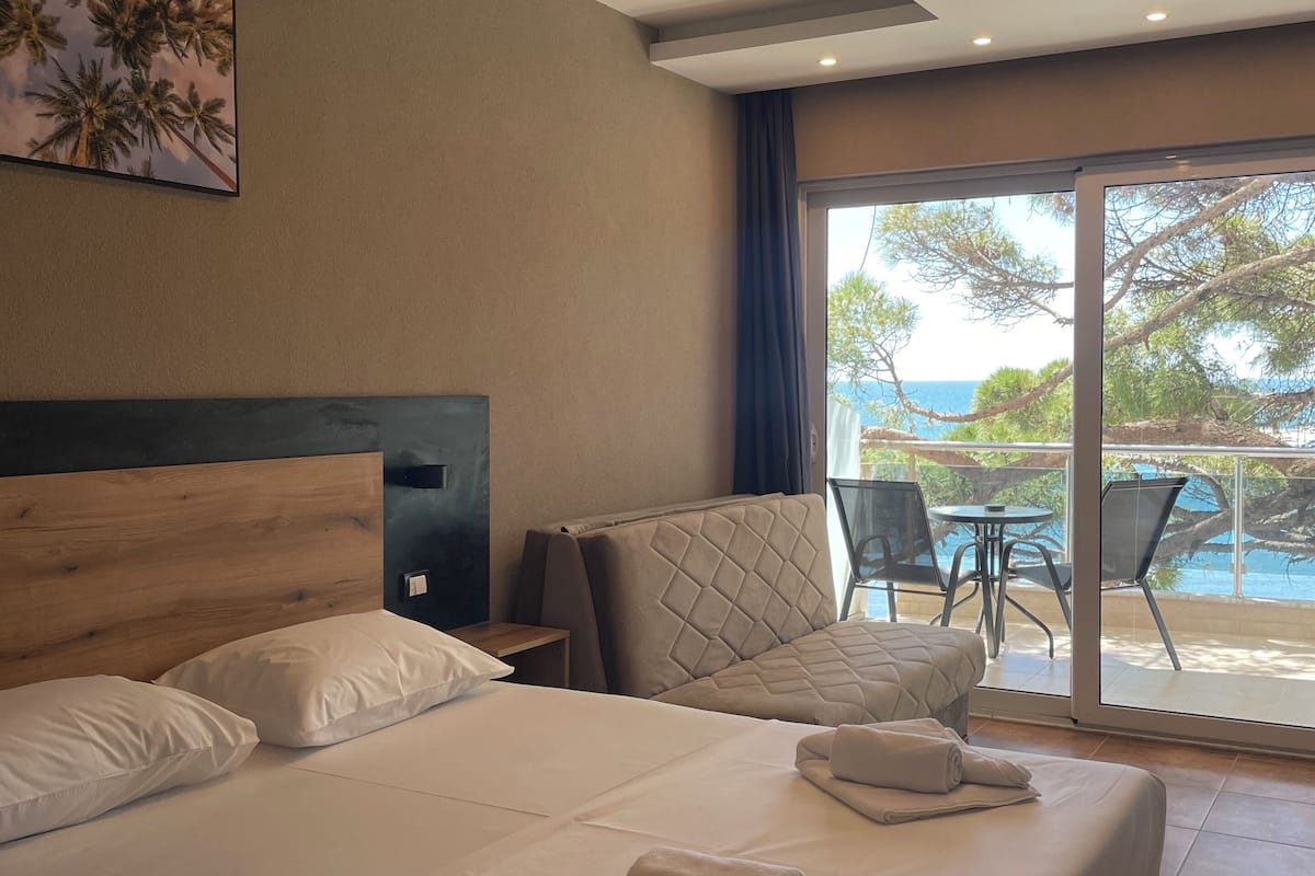 Una habitación de hotel con cama, cabecera, un cuadro, ventana con vista al mar y balcón con sillas.