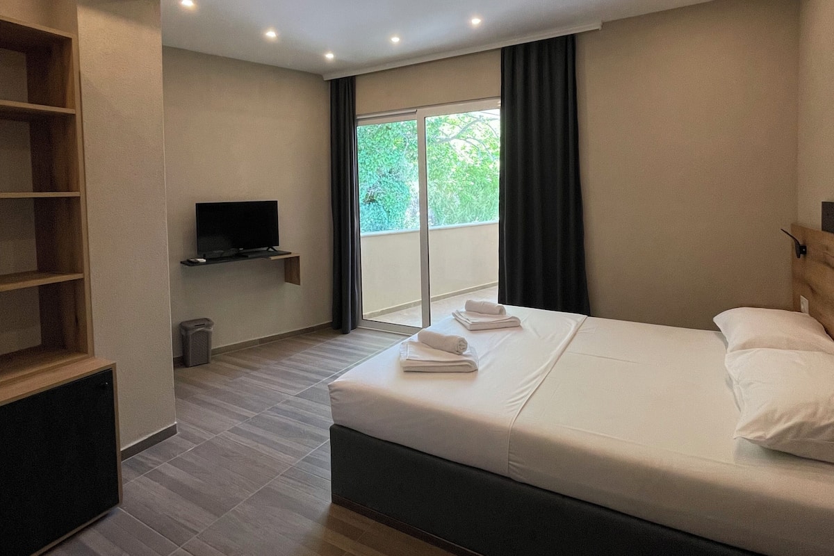 Habitación de hotel moderna con una cama grande, televisión, estante de madera y balcón con vistas.