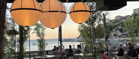 Un área de comedor al aire libre con mesas y sillas, lámparas colgantes grandes y vistas al mar y a los barcos.