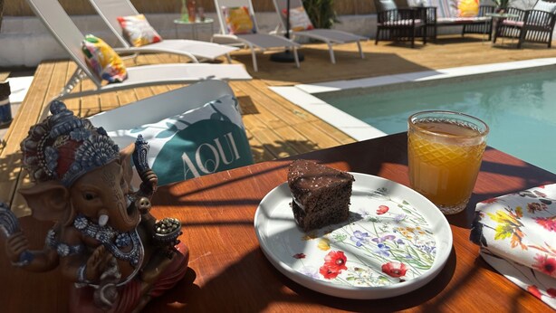 Une table placée au bord de la piscine, avec une part de gâteau, un verre de jus d’orange et une statuette d’éléphant décorative.