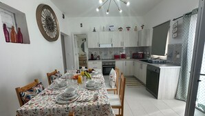 Une cuisine moderne avec des meubles blancs, une table à manger dressée pour quatre personnes et une grande horloge au mur.
