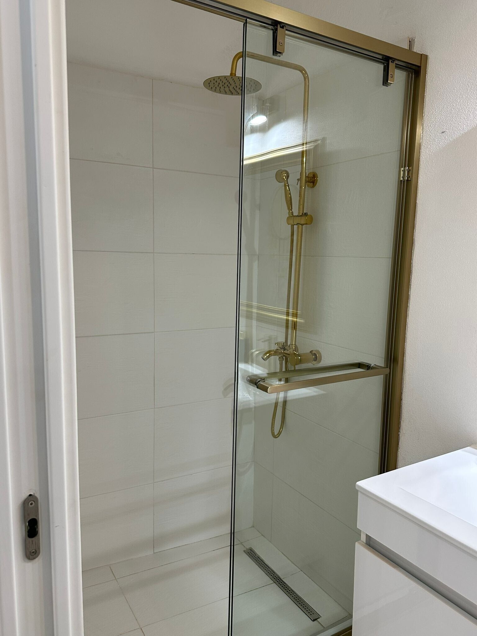 A glass-enclosed shower with a gold showerhead and handheld showerhead, a white tiled wall, and a white sink with a countertop.