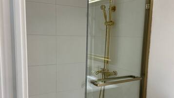 A glass-enclosed shower with a gold showerhead and handheld showerhead, a white tiled wall, and a white sink with a countertop.