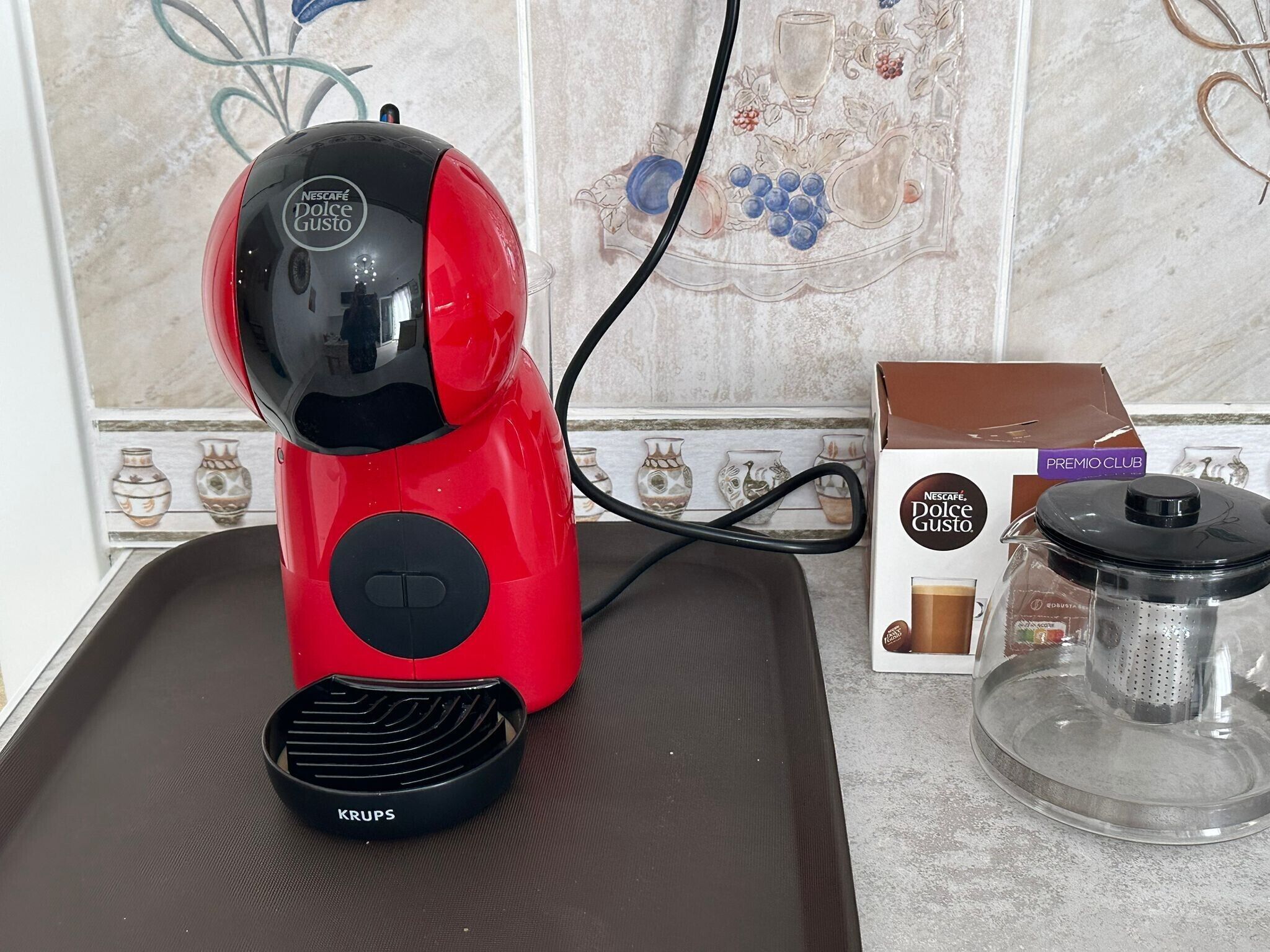 Punainen Krups -kahvinkeitin keittiön työtasolla, sen vieressä Nescafé Dolce Gusto -kahvikapselipaketti ja kahvipannu.