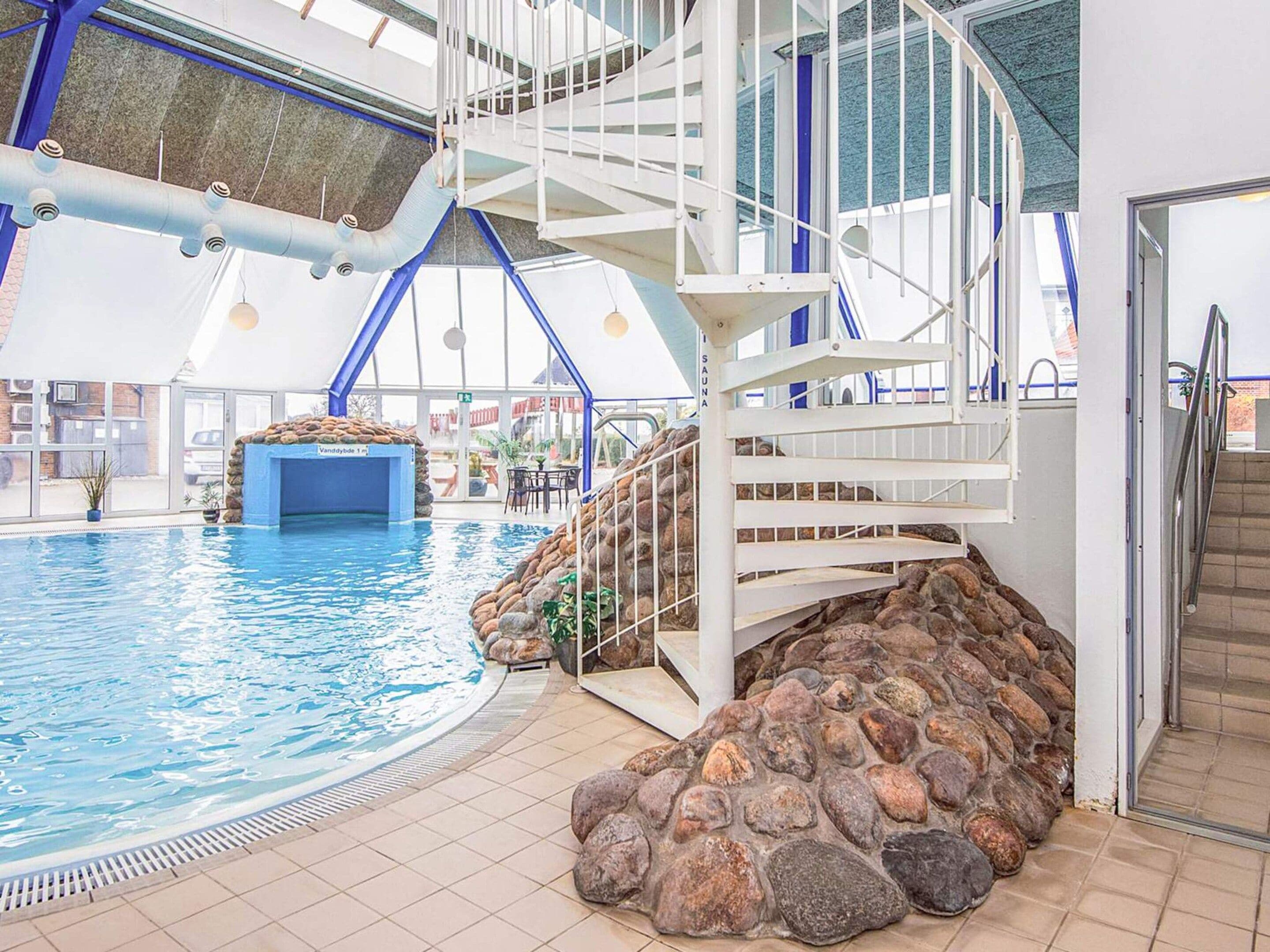 An indoor swimming pool with a stone wall and a white staircase leading to a roofed area.