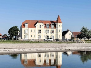 A large, multi-story building with a red roof, situated by a calm body of water reflecting its structure. - 4 Person Holiday Home in Hejls (Hejls)