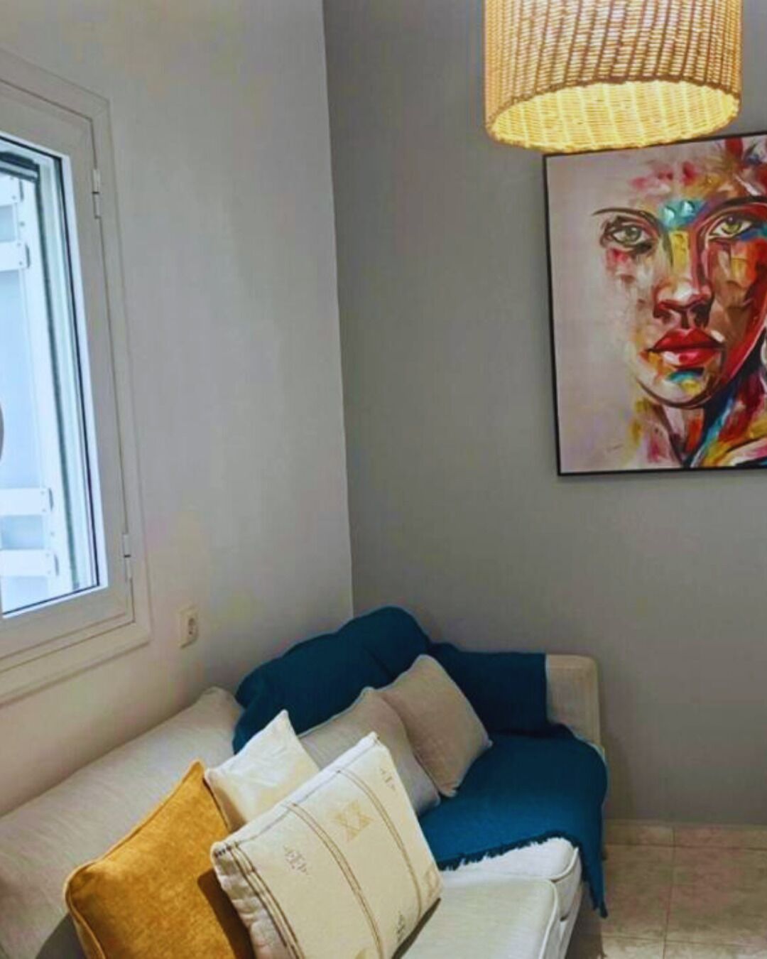 Una habitación con sofá, un cuadro, una ventana y una lámpara de fibra trenzada.