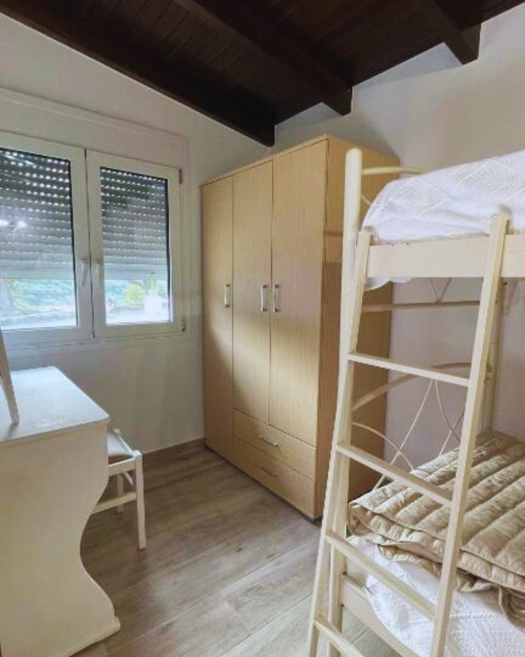 Une chambre avec un lit superposé, une armoire en bois, un bureau et une fenêtre avec des stores.