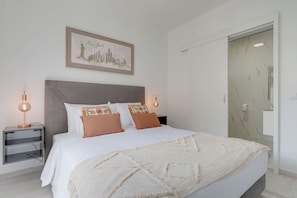 Un lit bien fait, avec une couette blanche et des coussins décoratifs, une œuvre d’art représentant un paysage urbain encadrée au-dessus du lit, une table de chevet avec une lampe, et une salle de bain avec un mur en marbre et un porte-serviettes.