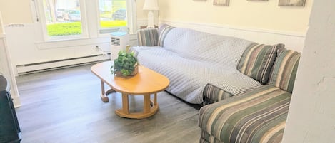 A living room with a sofa, a wooden coffee table, and a lamp. There are windows with a view of greenery outside.