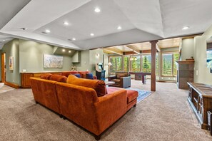 A spacious living area with a large orange sofa, a fireplace, and a view of a forest through large windows. - Alpenview Lodge | Best Of Breck (Breckenridge)