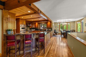 A kitchen with wooden beams, a central island with bar stools, a dining area with chairs, and a view through a window. - Alpenview Lodge | Best Of Breck (Breckenridge)