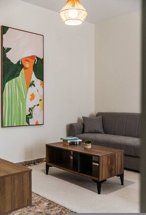 A living room with a grey sofa, a wooden coffee table, a framed abstract painting, and a hanging light fixture. - Perla Apartment 2BR Beachside (Golem)