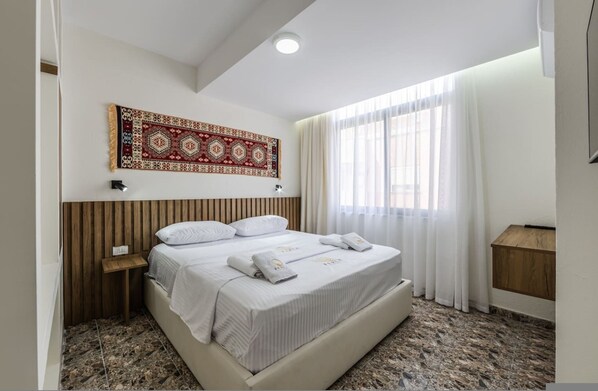 A hotel room with a bed, a nightstand, a wall-mounted tapestry, a window with curtains, and a wooden cabinet. - Perla Apartment 2BR Beachside (Golem)