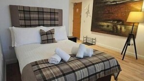 Una cama bien tendida con colcha a cuadros y almohadas, una lámpara de pie y un cuadro en la pared.
