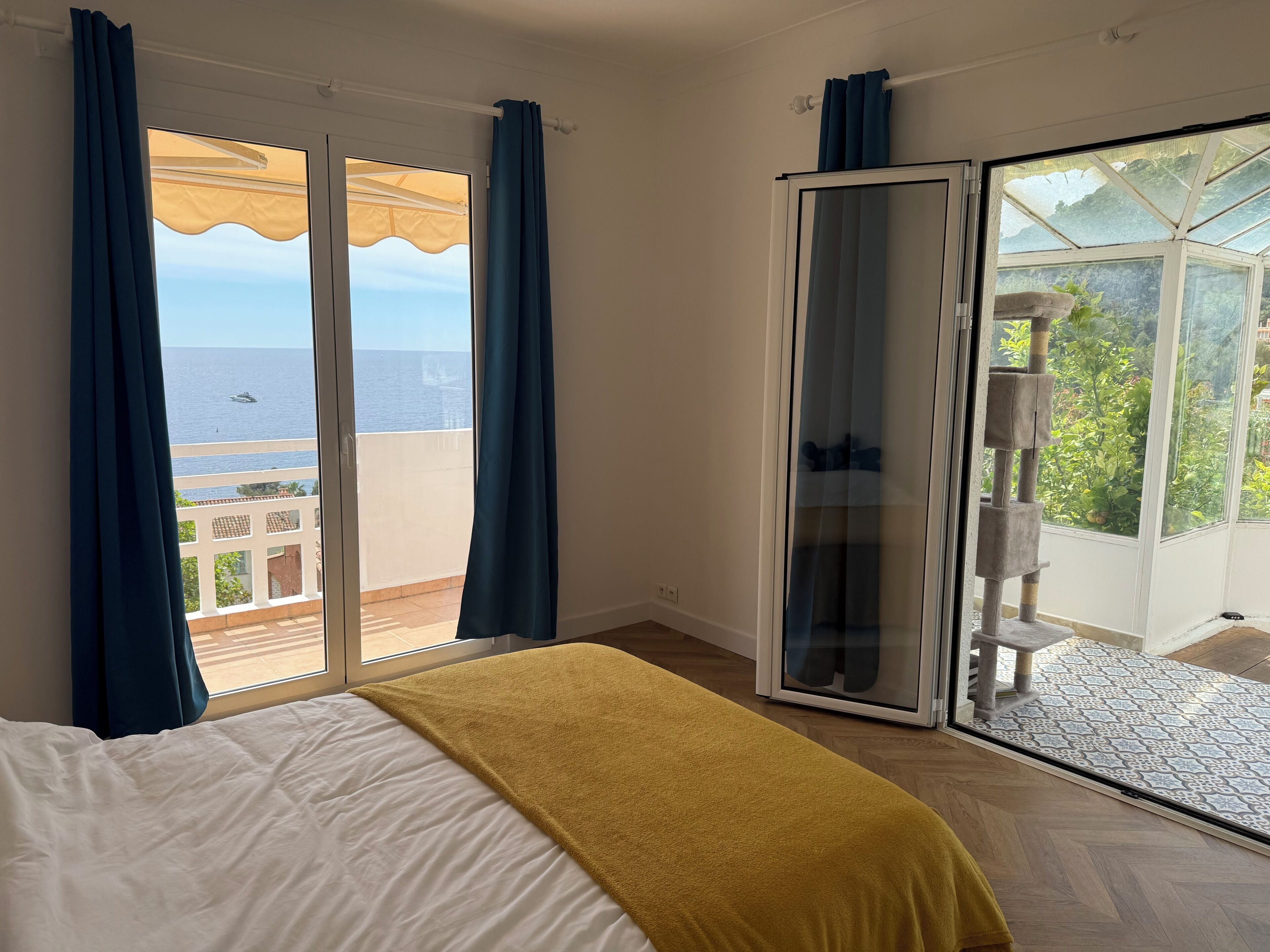 A bedroom with a bed, a yellow throw, a sliding glass door to a balcony, and a view of the sea.