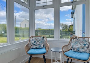 A room with two wicker chairs with blue cushions, a small round table, and large windows offering a view of a grassy area and a building.