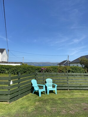 A grassy outdoor area with two turquoise Adirondack chairs, a wooden fence, and a view of houses and hills in the distance.