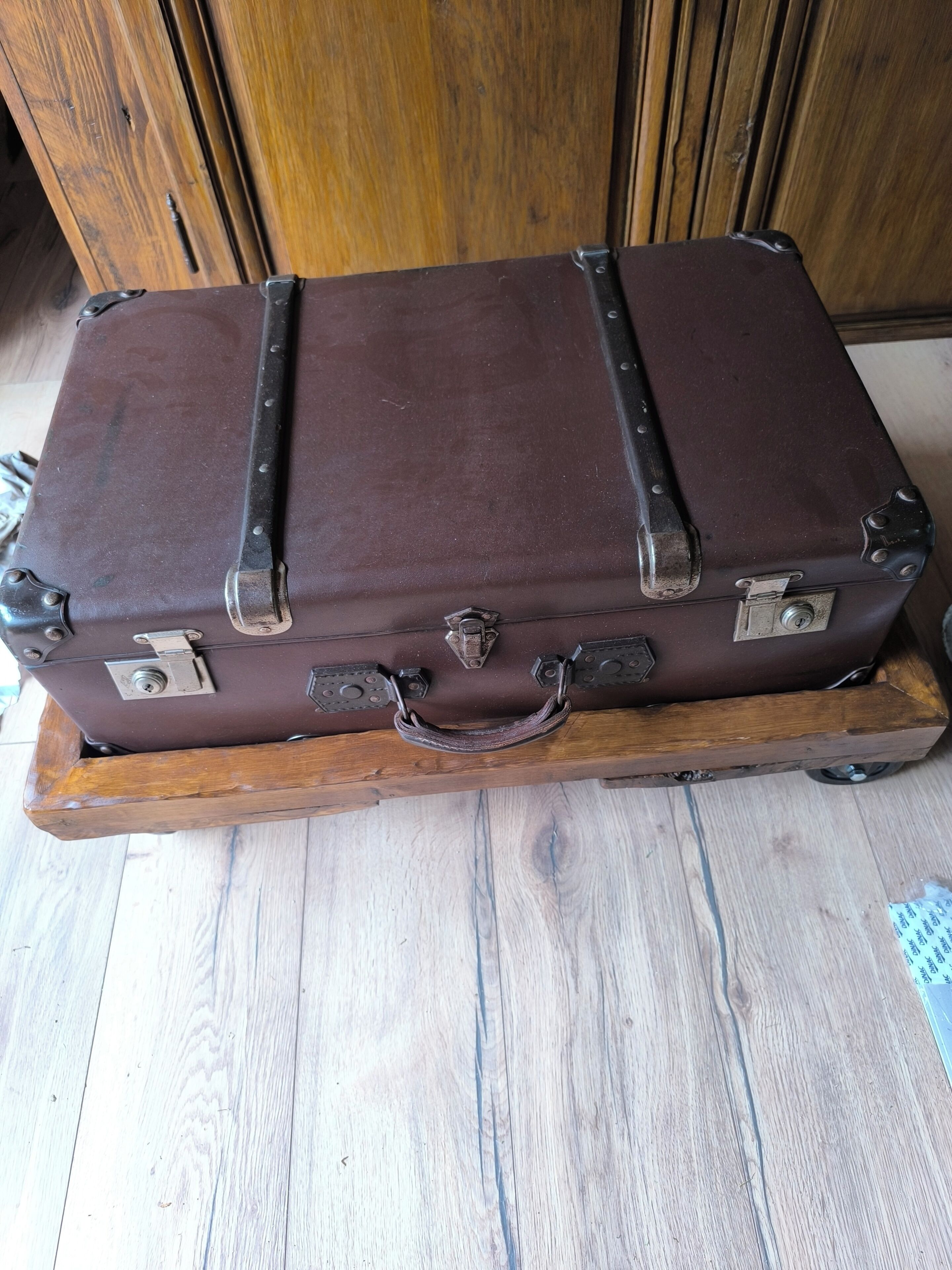Une valise marron avec des fermoirs métalliques et une poignée, posée sur un chariot en bois.
