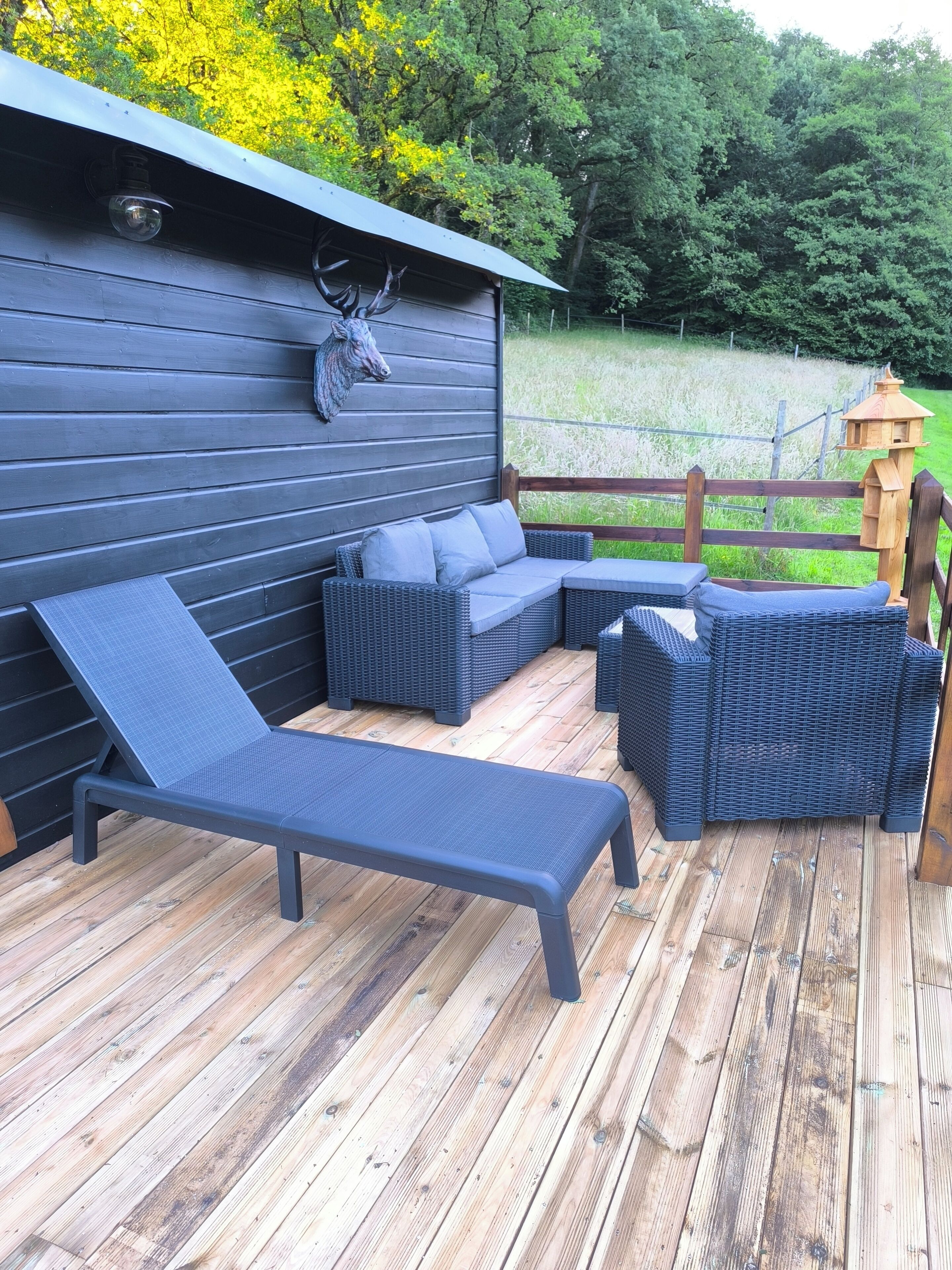 Eine Terrasse mit einer dunkelgrauen Couch, einem Liegestuhl und einem Holzzaun.