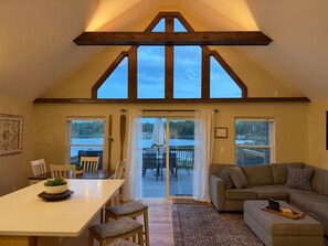 A spacious room with a high ceiling, wooden beams, a dining area, a sofa, and large windows overlooking a body of water.