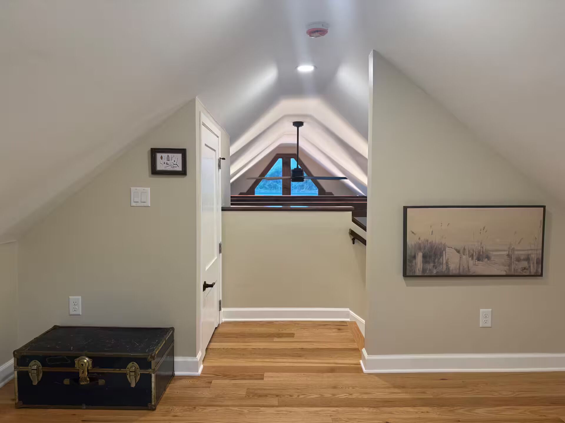 A spacious attic room with a wooden floor, a large window, and a ceiling fan.