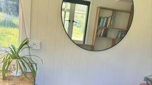 A room with a round mirror, a wooden table, two cushioned seating areas, a potted plant, and a bookshelf.