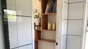 A corner of a room with white tiled walls, a wooden bookshelf with books and candles, and a potted plant.