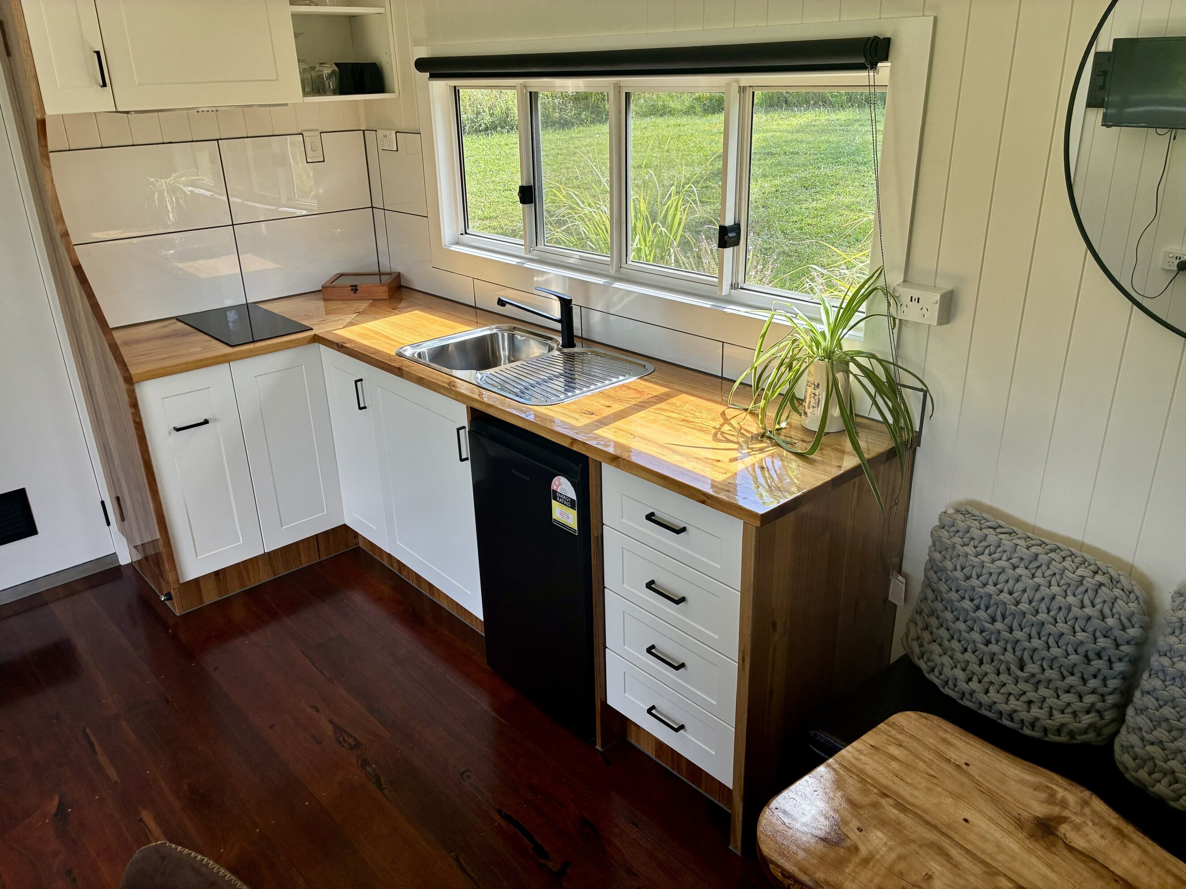 A compact kitchen with a sink, wooden countertop, white cabinets, and a window overlooking a grassy area.