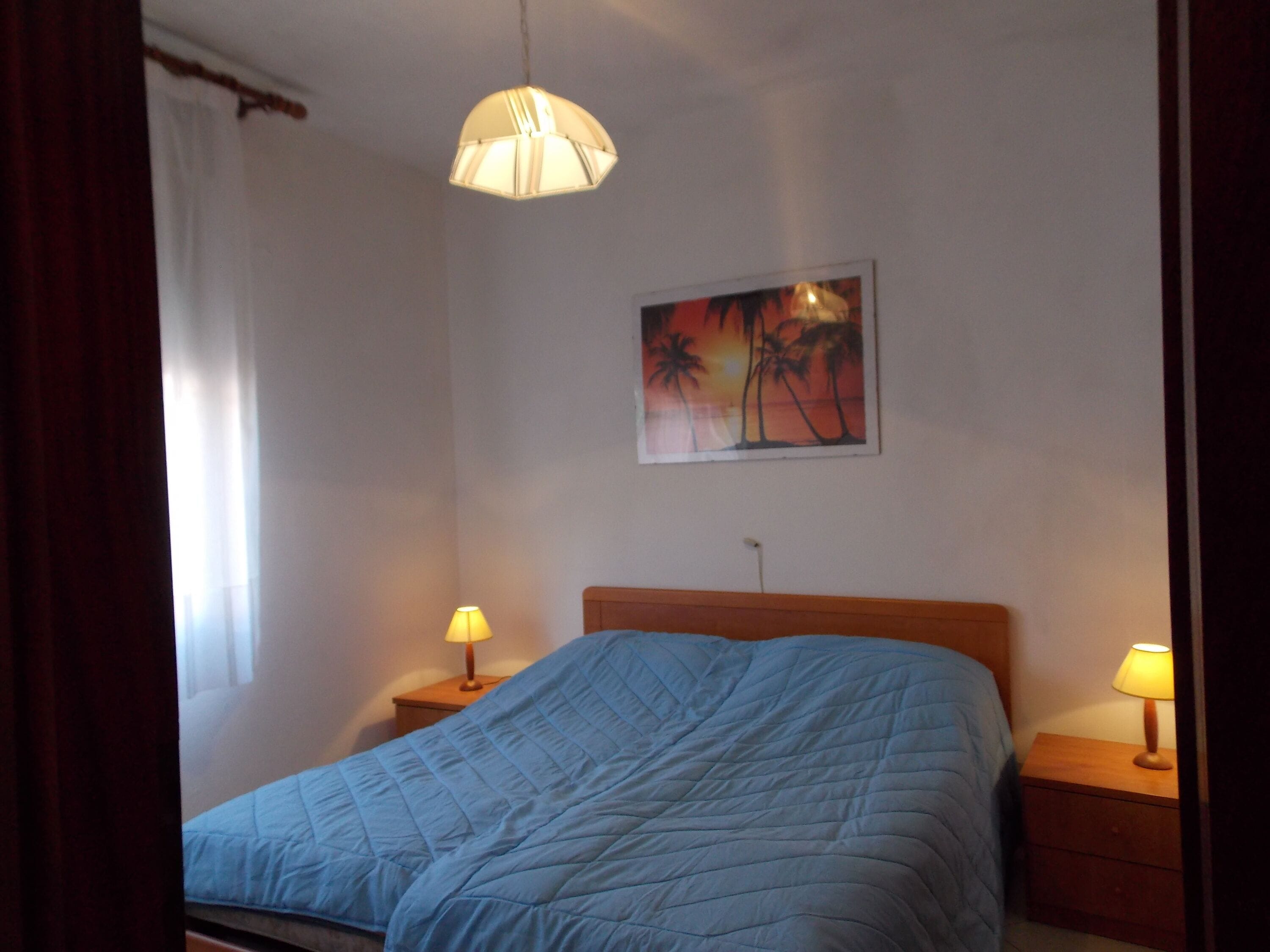 Une chambre à coucher avec un lit, deux tables de chevet avec des lampes, un tableau au mur et une fenêtre avec des rideaux.